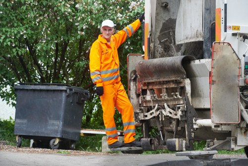 A waste collection worker dressed in bright orange high-visibility safety clothing, including a jacket and trousers with reflective stripes, is standing on a residential street near a large industrial rubbish compactor vehicle. The worker is holding a black wheeled rubbish bin positioned beside the vehicle, which has an open hopper at the front for waste loading. The scene takes place outdoors, with greenery and trees visible in the background, and the pavement beneath the worker and vehicle appears clean and well-maintained. The vehicle is equipped with various mechanical parts and safety features, indicating it is designed for efficient rubbish collection and disposal. The overall setting reflects a typical rubbish removal operation possibly carried out by Business Waste Removal Harrow in the Harrow area, with the worker preparing to load waste as part of routine waste management services, situated on a tidy street surface that relies on professional waste removal support to keep the local environment clean.