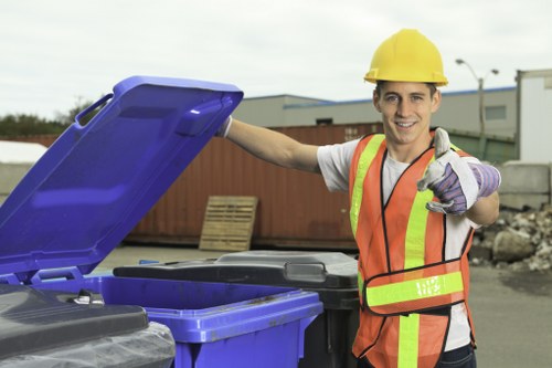 A young male waste management worker wearing a yellow safety helmet, with a bright orange high-visibility vest over a light grey t-shirt, standing outdoors near a row of black and blue rubbish bins on a paved area. He is smiling and giving a thumbs-up while holding the lid of the blue bin open. The background includes stacks of wooden pallets, a corrugated metal fence, and industrial buildings under a cloudy sky. The scene illustrates waste collection and rubbish removal services, with the worker actively engaged and prepared for rubbish disposal tasks typically associated with locality near Harrow and its surrounding areas. The environment appears clean and organized, emphasizing professional waste handling in an urban or suburban setting.